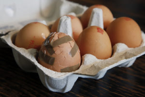 A photograph of brown eggs in an egg carton. One egg is cracked and broken. It has been taped together again.