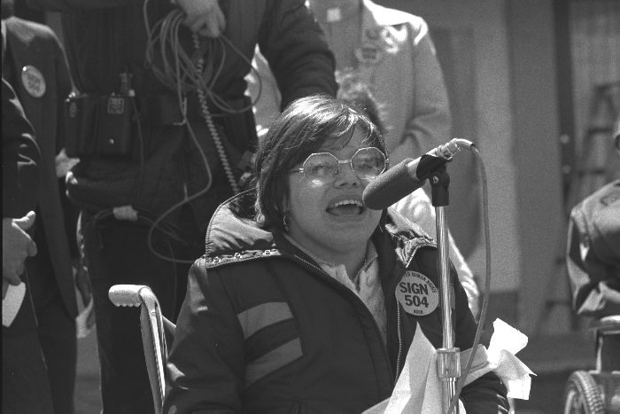 A black and white image of a young woman sitting in a wheelchair, Judy Heumann, speaking into a microphone at a demonstration.