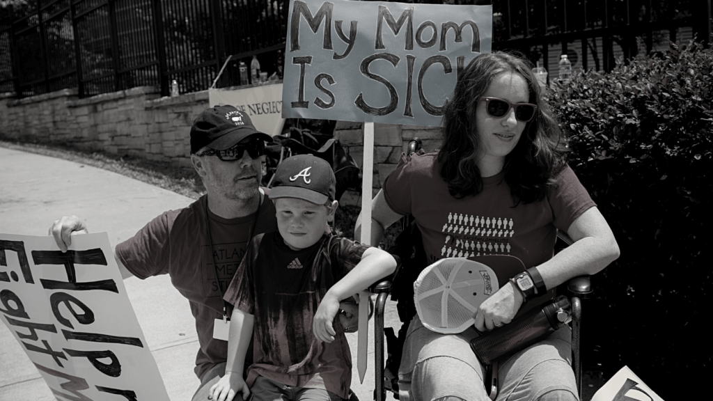 father, son and mother in wheelchair protesting outside CDC. Signs read "My Mom Is Sick" and "Help me fight ME"