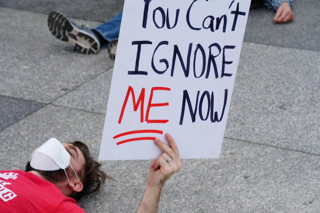 Ben laying on the cement in a red shirt and white facial mask. Holding a protest sign that says "You Can't Ignore ME Now"