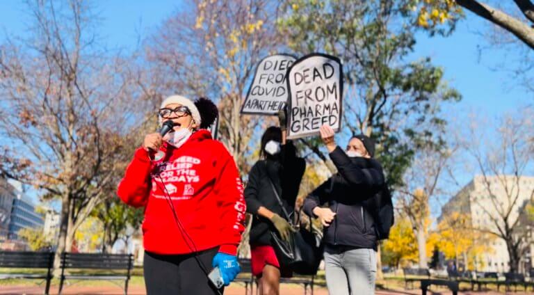 Blue sky with trees with some foliage and others bare in a park with benches and office builds on the right and left sides. In front stands stands a light skinned black woman in black framed glasses, a red hoodie and a white beanie holding a microphone in one hand with her mouth open wide speaking. Behind her is a dark-skinned black woman with shoulder length hair in a white mask, black top and red skirt with a black purse across her shoulder. Above her head she holds a sign painted gray and shaped as a gravestone with black lettering that reads "Died from COVID apartheid." To the right is a white protestor in a puffy black coat with gray jeans and a black hat holds another gravestone sign that reads "Died from Pharma Greed."