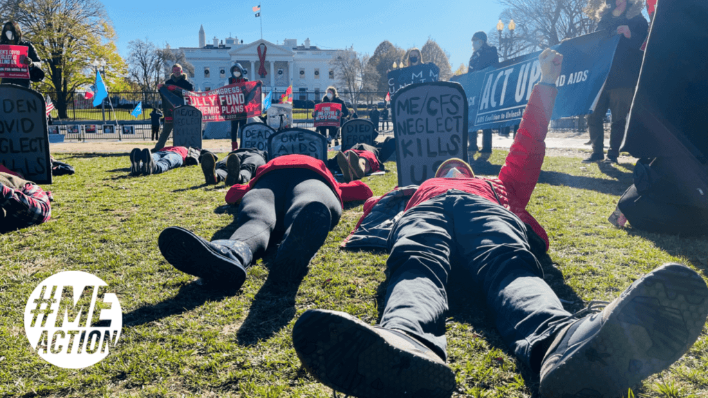 A protestors lie on the grass in park in front of the White House, on which a giant AIDS ribbon hangs. Protestors standing in the background hold signs saying "Biden and Congress: fully fund pandemic plans" and "ACT UP, Fight AIDS." Protestors lying on grass hold painted gravestones at their heads. Man lying on ground in mask closest to camera wearing dark pants and a red shirt, orange coat, and yellow beanie with white mask. His holds aloft his left fist clenched and holds behind him a gravestone that reads "ME/CFS neglect kills us." A ray of light from the sun streaks the right side of the image. On the left is a logo of a white circle with transparent letters "#MEAction."
