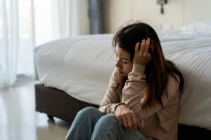 Young woman sits on floor at edge of bed looking distraught with head in hand