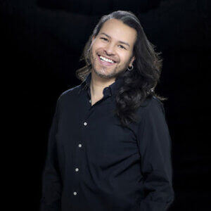 Gabriel is wearing a black shirt with long brown hair and is smiling.