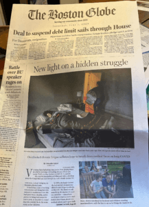 Screenshot of a newspaper showing the front page of the Boston Globe. There is a large photo above the fold showing a woman lying in bed with her husband in the doorway, and a second photo showing a family roasting marshmallows in their backyard, with the mother at a table in the distance.