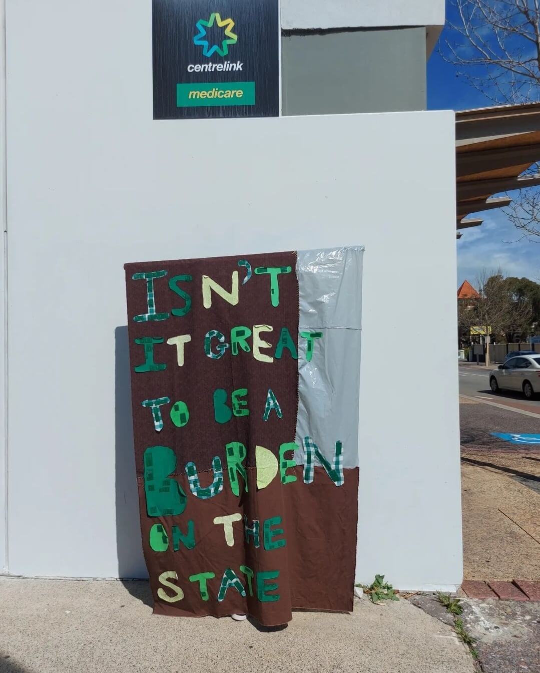 Sign written in green and yellow that reads, “Isn’t it great to be a burden on the state”.