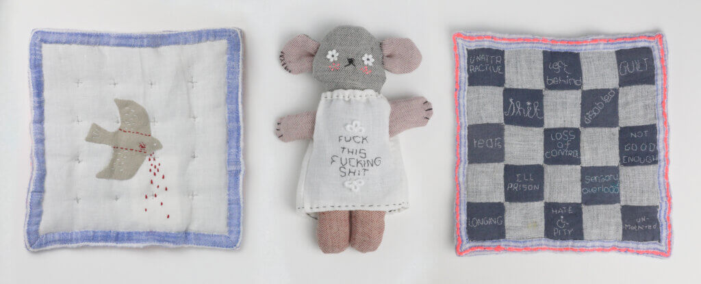Cloth of a bird with blood coming out of it. Beside the cloth is a teddy bear wearing shirt that reads, “Fuck this fucking shit”. Beside the bear is a checkered cloth with negative feelings written on black squares.