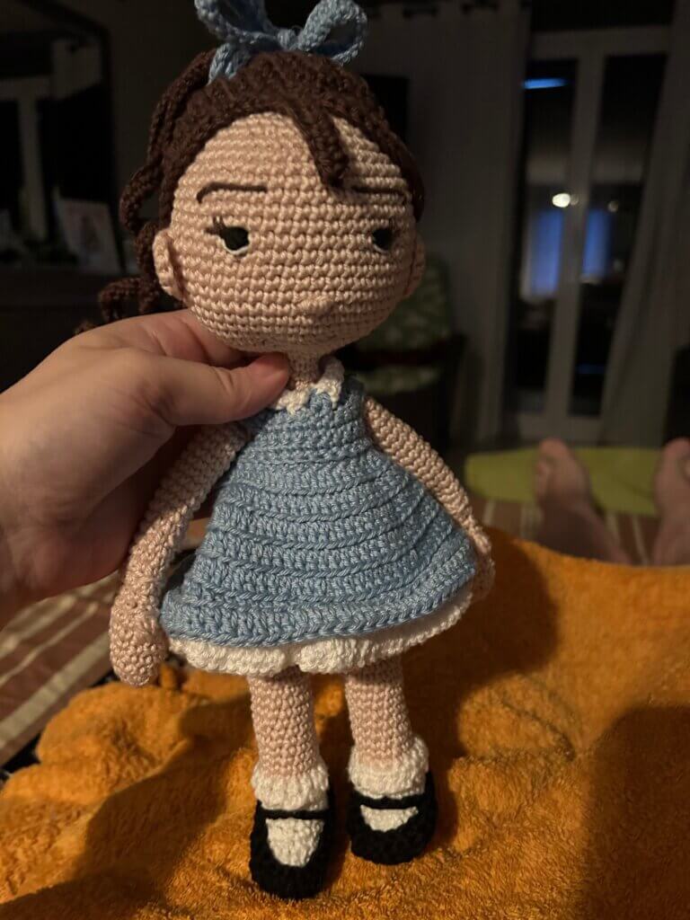 A crocheted doll of a little girl wearing a blue dress, black Mary jane shoes with white riffled socks, and a blue bow in her brown hair
