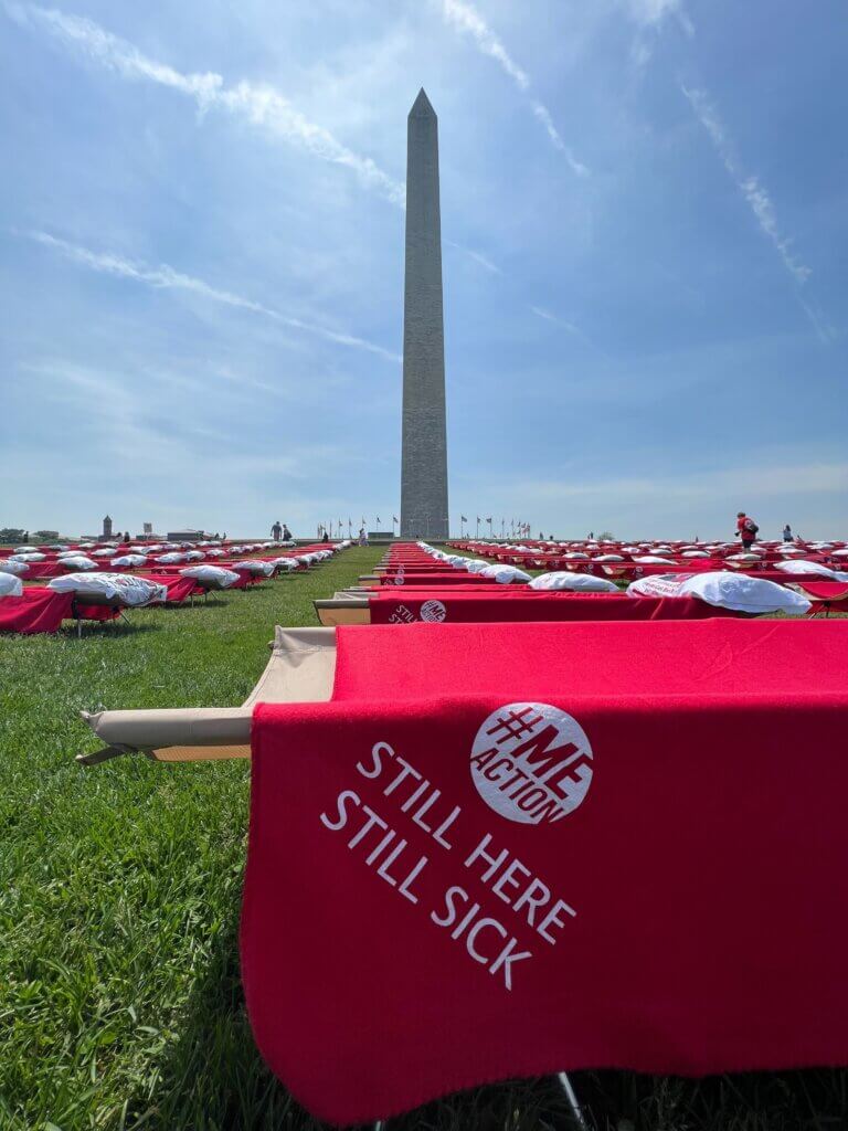 red blanked with the words still here, still sick and the #MEAction logo on it. the blanket is laying over a cot with other cots on a grassy field in front of the Washington Monument.
