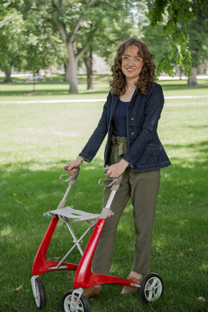 A white adult woman is standing holding onto a vibrant red futuristic-looking rollator. She is smiling and is in a grassy park with trees, wearing wavy curly brown hair, pink glasses, and a dark denim jacket with olive green high-waisted pants.