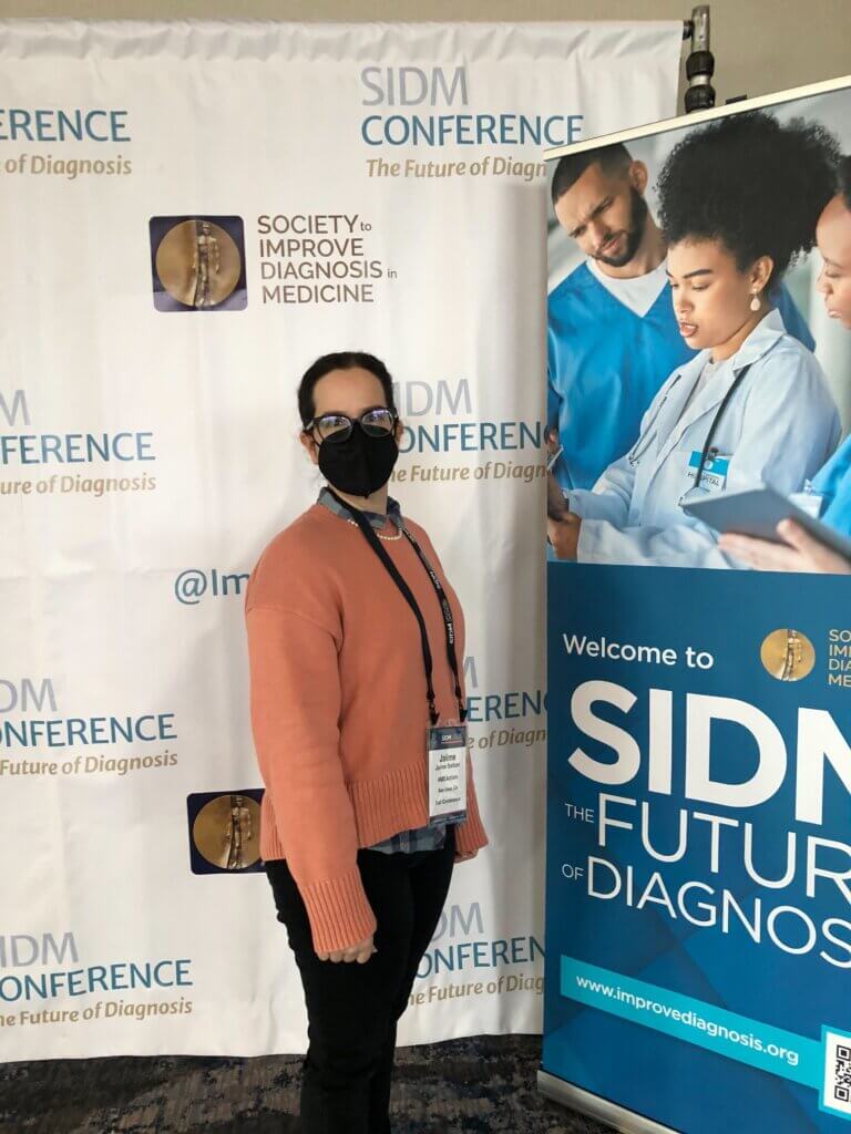 Jaime Seltzer stands in front of a SIDM conference poster backdrop, wearing a pink sweater with a button-down underneath.  She is a white woman with pinned-back dark hair, and is wearing black glasses and a black face mask.