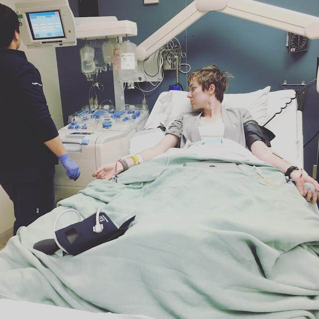 A white adult woman lays in a reclined hospital bed with arms outstretched. There is a blood pressure cuff attached on one arm, an IV placed in both arms with medical tubing connecting to a complex medical machine on her left for blood filtering. A medical tech stands at the medical machine.