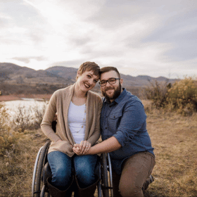 A portrait style couple photo with Alison and her husband in the same location and same clothing as the previous photo, but Alison is seated in her wheelchair and smiling at the camera while her husband kneels next to her, holding her hand and posing for the camera.