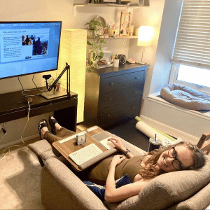 A bird’s eye view of a home office, with Alison, a white adult woman laying in a recliner with a lapdesk containing keyboard and trackpad on her lap. There is a mounted TV screen on the wall in front of her as computer display. She is looking at the camera, smiling, wearing black glasses, olive green dress, and wavy brown hair.