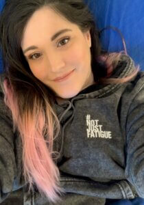 Elizabeth is lying in bed smiling at the camera. Her brown and pink hair rests over her right shoulder. She’s wearing a grey, acid wash #NotJustFatigue hoodie.
