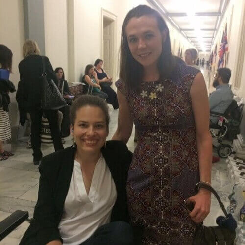 image of two women people in a hallway. One woman is standing and another woman is in a wheel chair. Both are smiling at the camera.