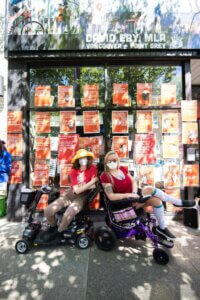 two people in scooters/wheel chairs in red shirts wearing masks. they are in front of a wall covered in red posters about me. there are shadows from a tree in the photo.