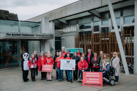 A group of people stand outside the Scottish Parliament building at Millions Missing Scotland 2022 looking serious. Many of them are wearing red ME Action t-shirts. Some people are holding signs that say Millions Missing. In the middle, someone is holding a large piece of card with a pledge on it and signatures of MSPs. One person is in a wheelchair and one is on a mobility scooter.