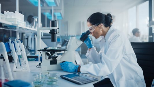 The image is of a scientific lab with a woman in a white lab coat looking through a microscope